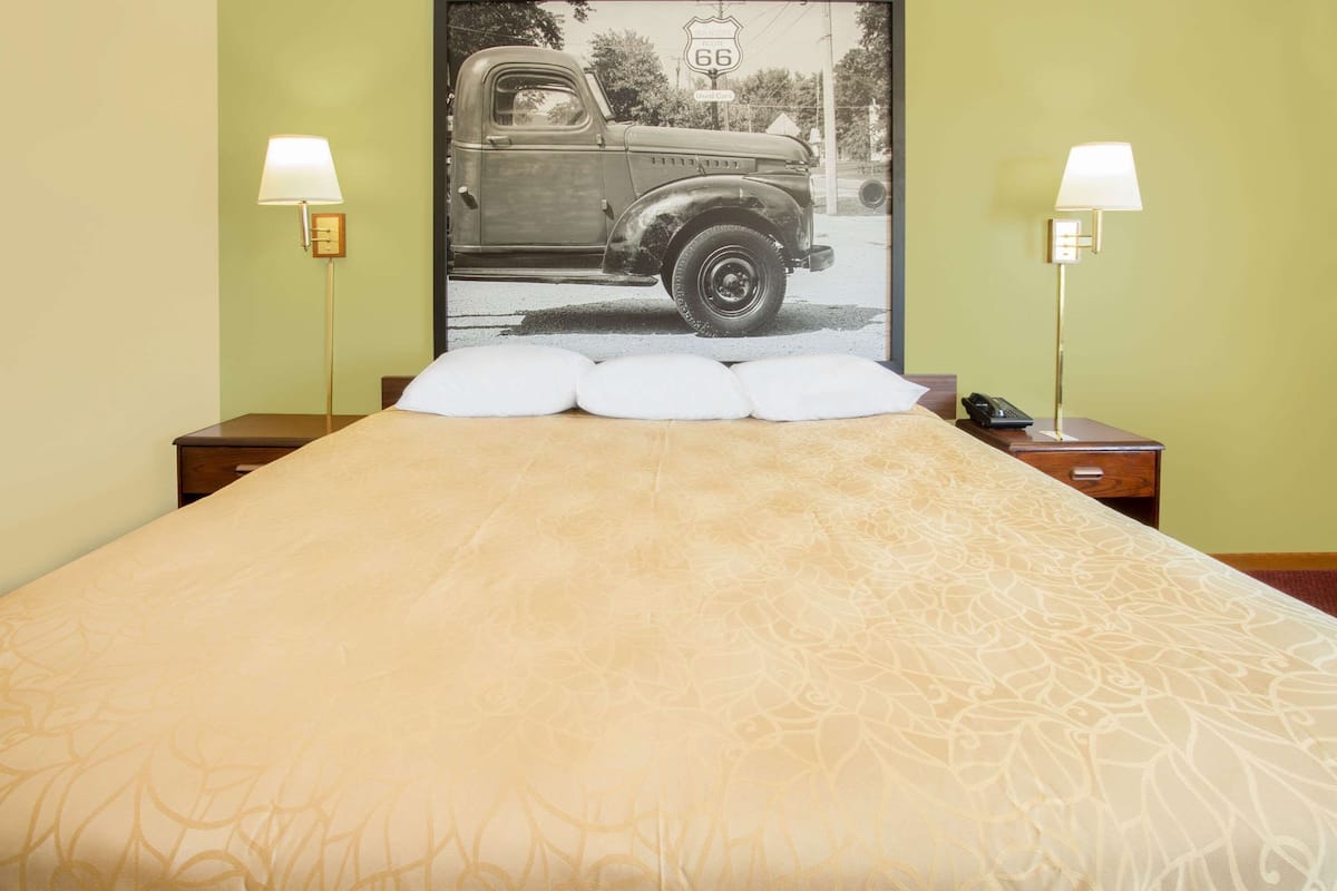 A bed with a patterned bedspread, two bedside lamps, a nightstand with a phone, and a poster of a vintage car on the wall.