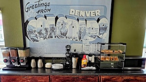 A counter with a large framed sign reading "Greetings from Denver" and "State Capital in Denver," coffee machines, a coffee dispenser, and a display case with pastries.