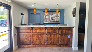 A reception area with a wooden counter, a framed welcome sign, and a wheelchair-accessible sign. - Deluxe Inn Willows (Willows)