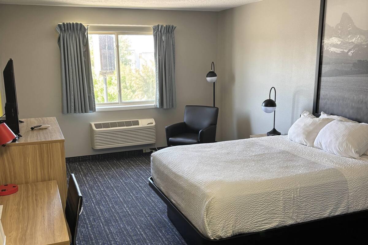Une chambre d’hôtel comprenant un lit, un bureau, une chaise, une fenêtre avec des rideaux, un climatiseur et une lampe.