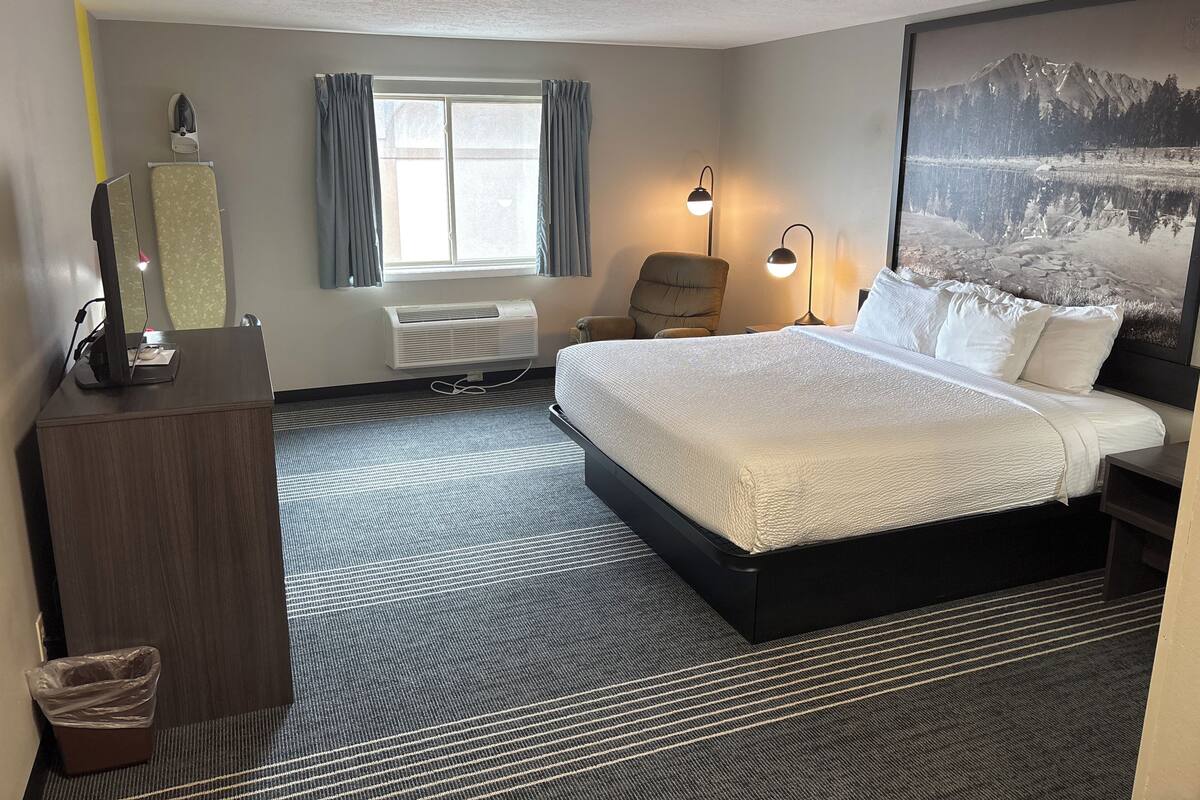 Une chambre d’hôtel avec un grand lit, un bureau avec un ordinateur, une chaise, une fenêtre avec des rideaux, un radiateur fixé au mur et un grand tableau encadré représentant un paysage de montagne.
