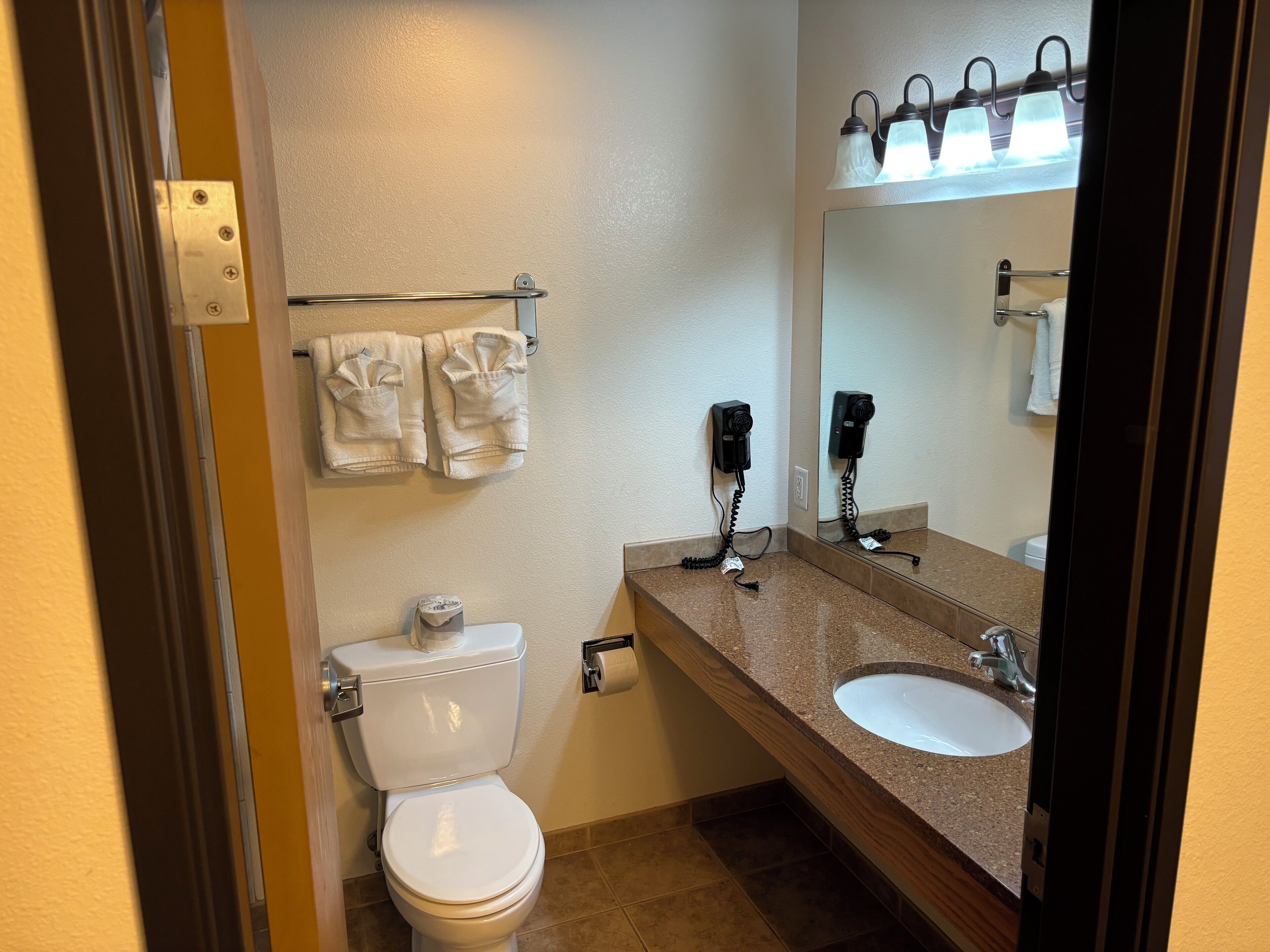 Un baño con inodoro, lavamanos y espejo. Hay toallas colgadas en un perchero y un secador de pelo en la pared.