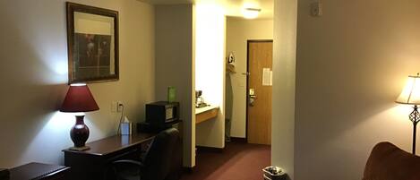 A hotel room with a bed, a desk with a computer, a chair, a lamp, and a door leading to another room.