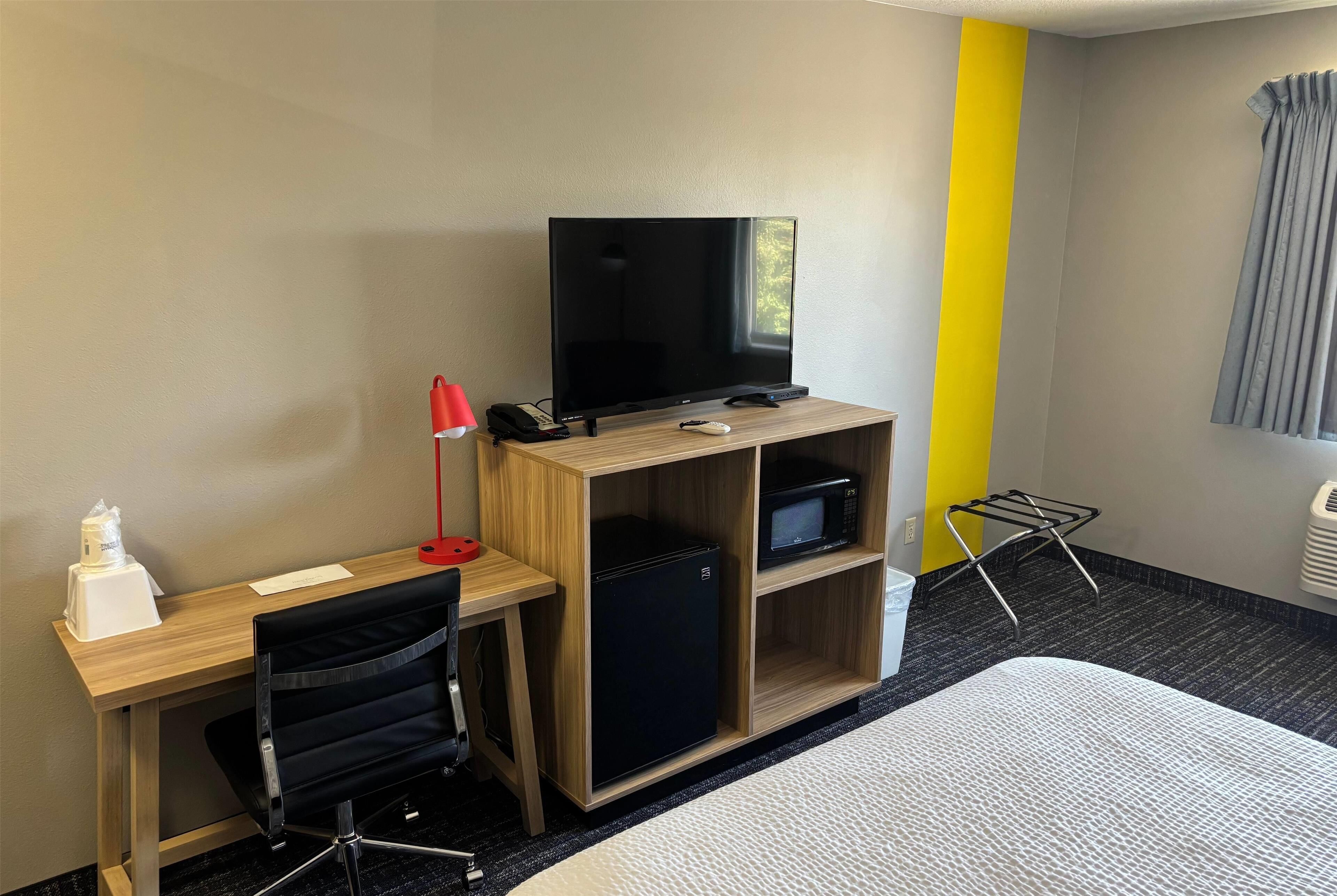 Une chambre d’hôtel avec un bureau en bois, une chaise noire, un téléviseur, un mini-réfrigérateur, un four à micro-ondes, une lampe rouge, une bouteille blanche et un lit recouvert d’une courtepointe à motifs.