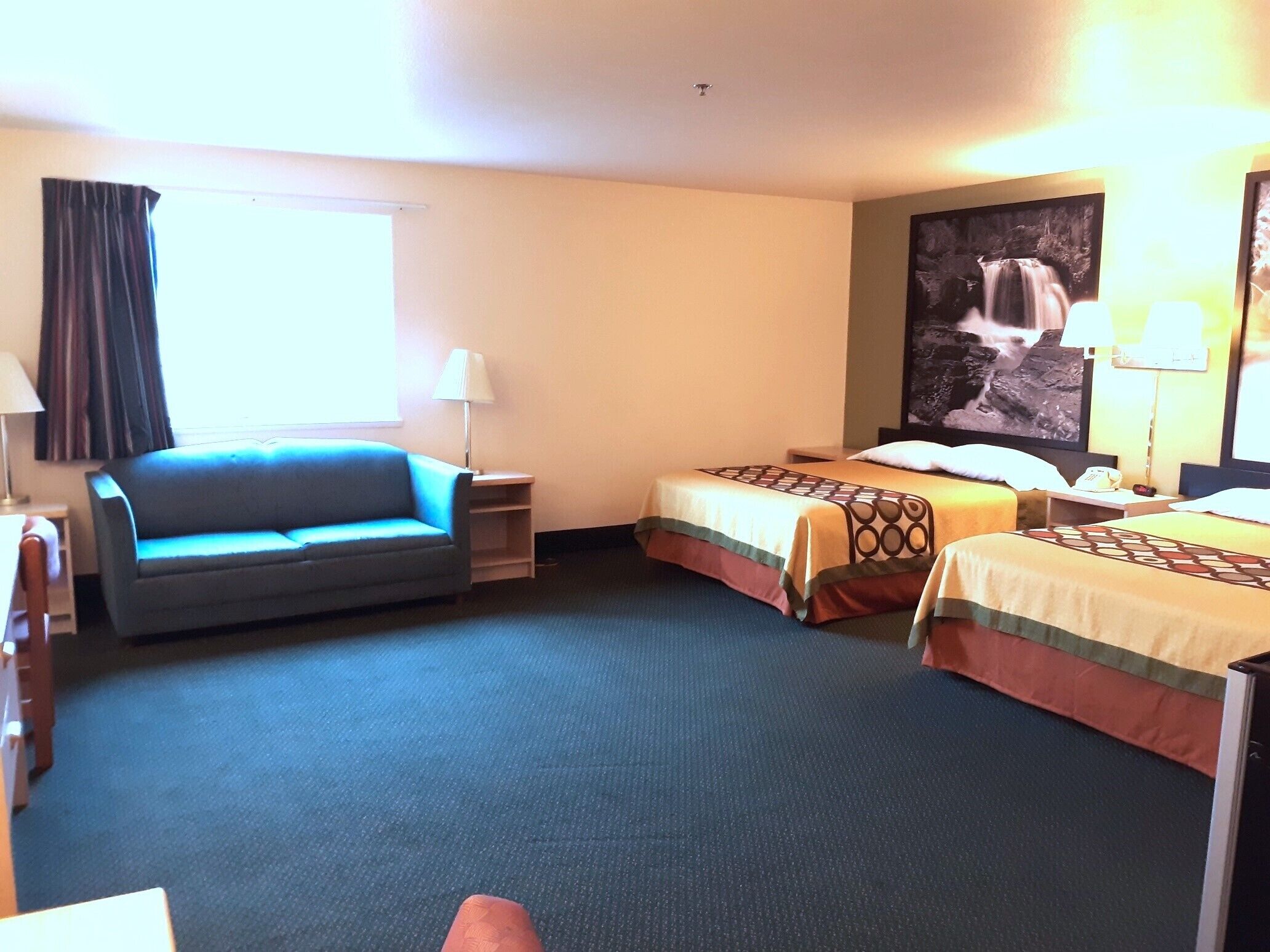 A hotel room with a sofa, two single beds, a desk, a lamp, and a large painting on the wall.