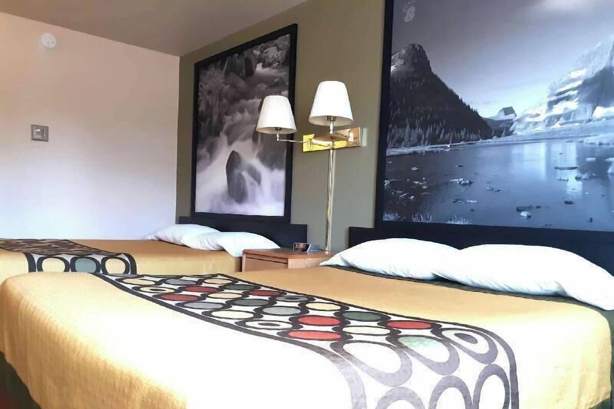 A hotel room with two beds, a large painting of a mountain landscape, a floor lamp, and a view of a body of water.