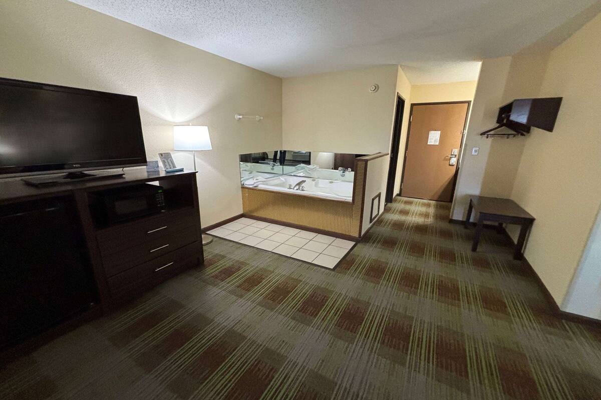 A hotel room with a flat-screen TV, a desk with a lamp, a bathroom with a sink and mirror, and a carpeted floor.