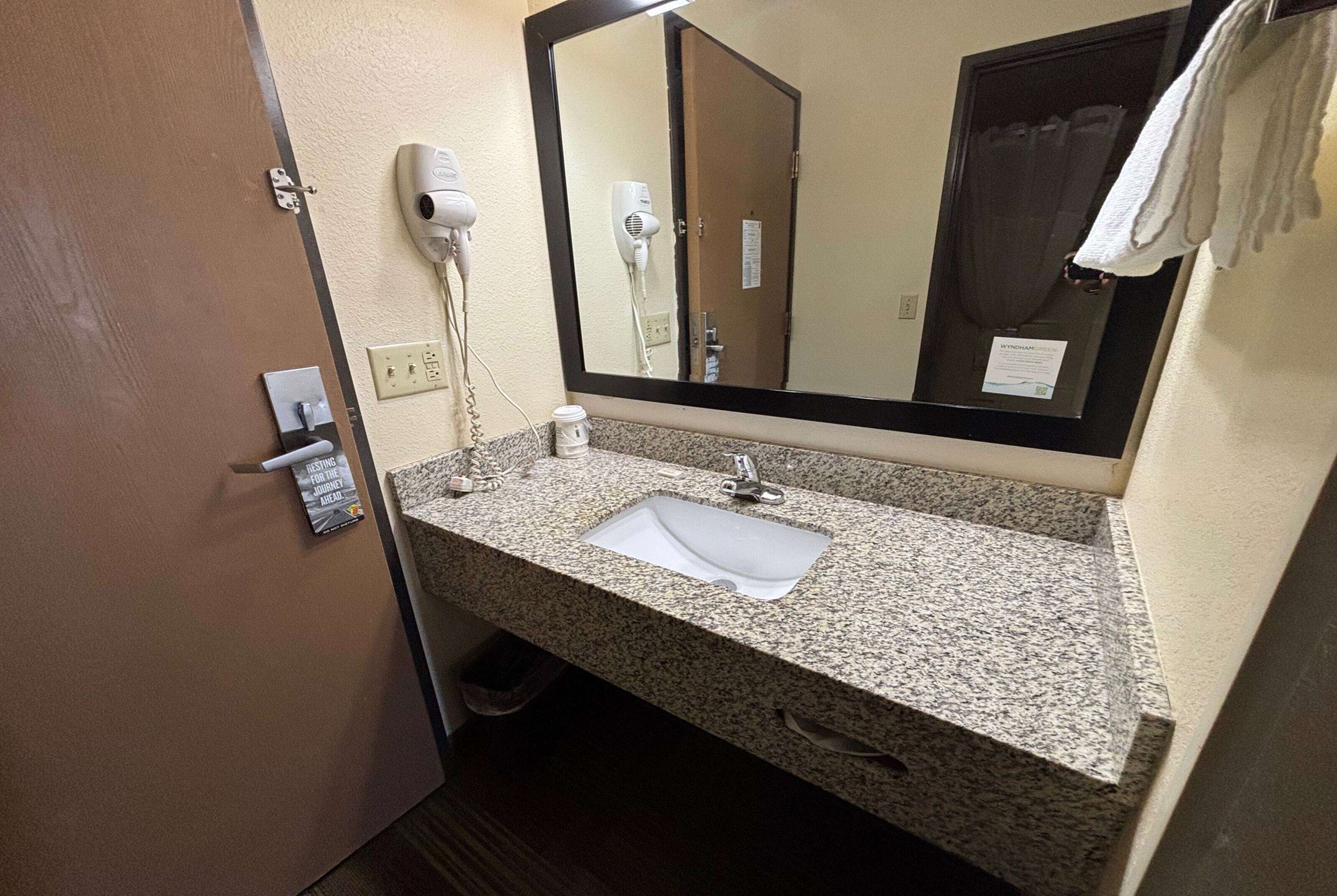 A bathroom with a granite countertop, a sink, a mirror, a hairdryer, and a towel hanging on the wall.