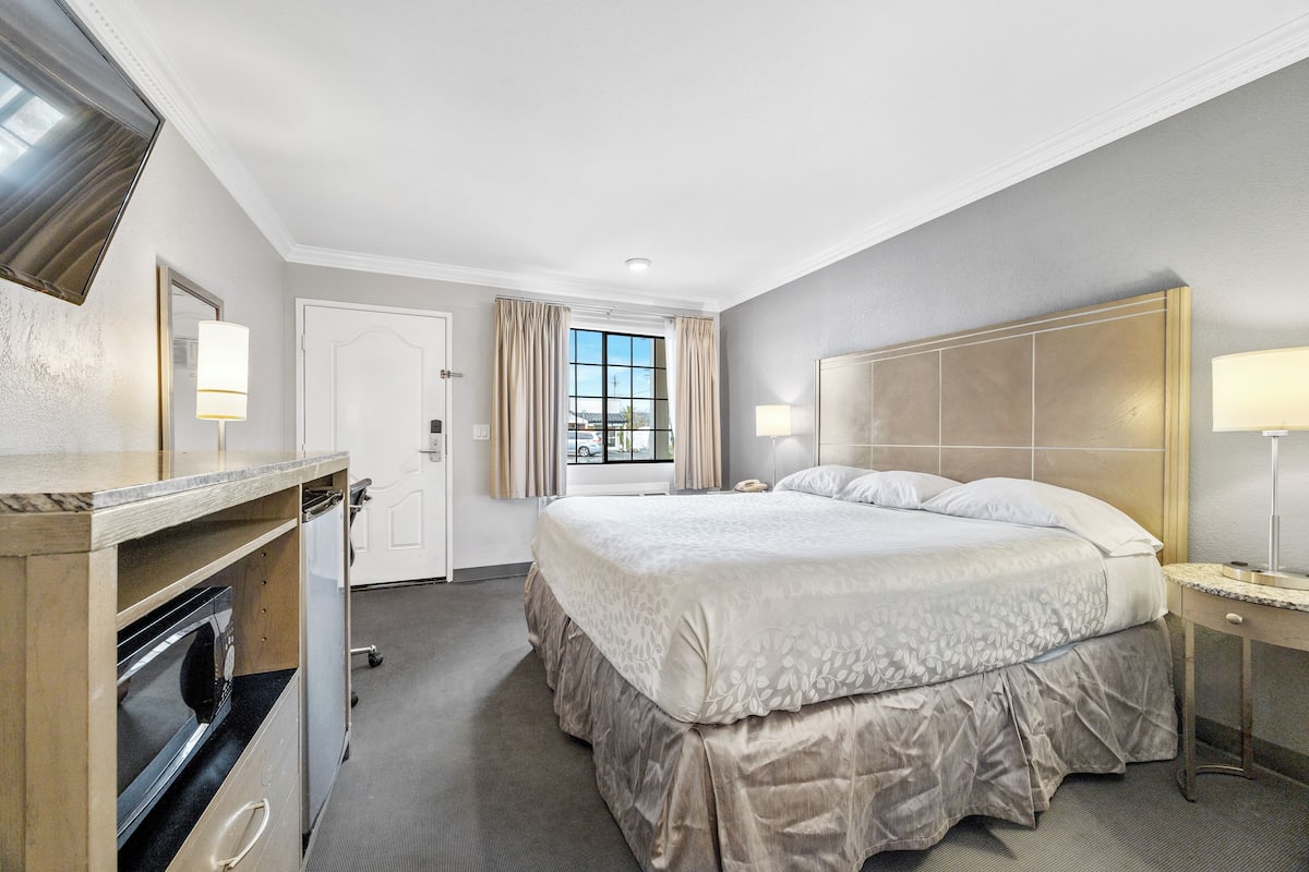 A hotel room with a bed, a desk, a chair, a lamp, a mirror, a door, and a window.