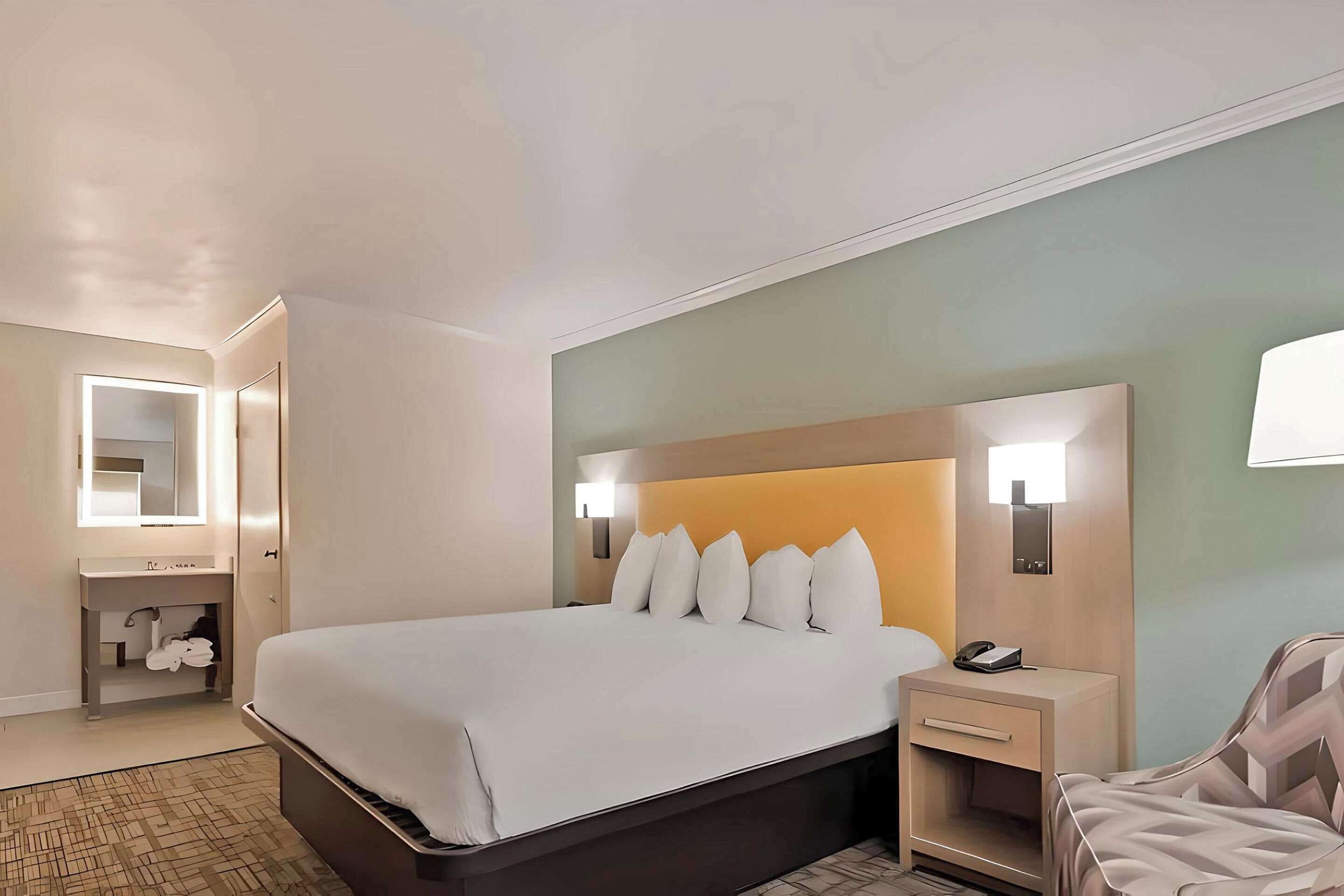 Una habitación de hotel con una cama grande, mesitas de noche, un espejo y un baño con lavamanos y toallero.