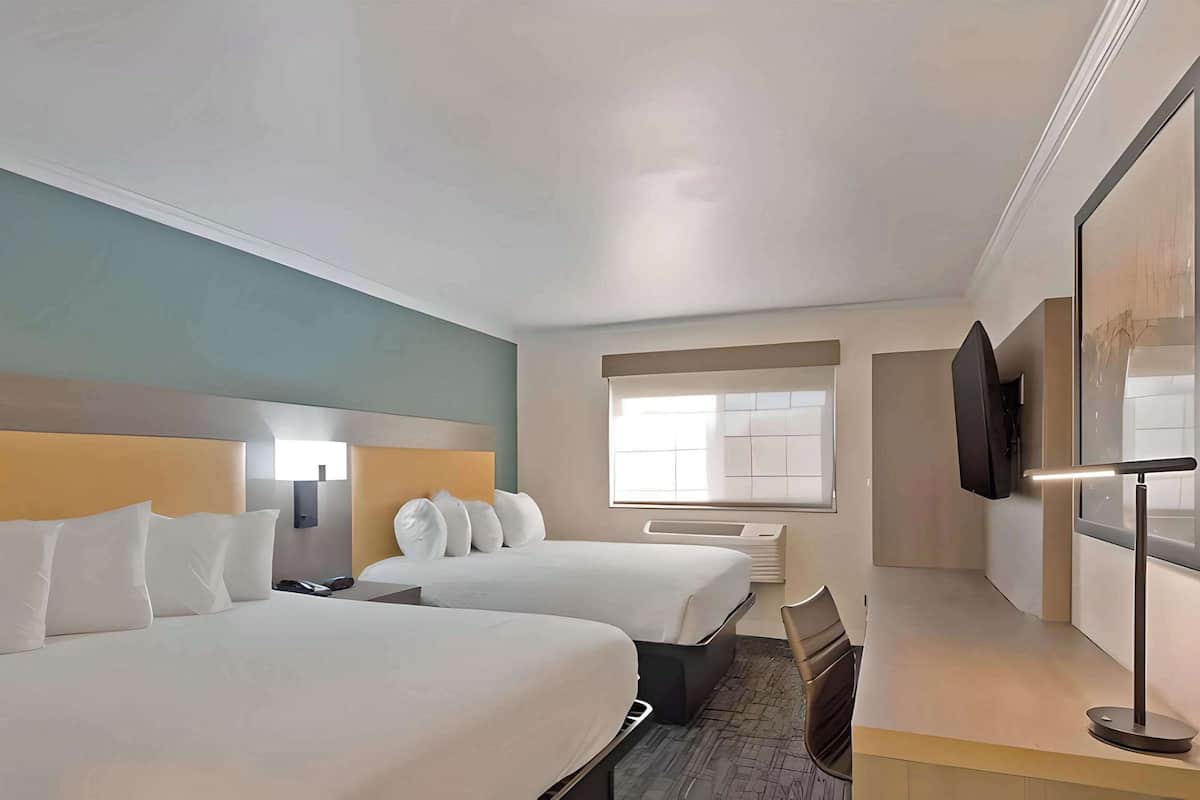 Quarto de hotel com duas camas, uma escrivaninha, uma cadeira, uma televisão e uma janela.