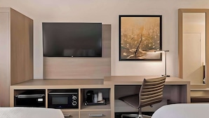 A modern hotel room with a flat-screen TV mounted on the wall, a desk with a chair, a bookshelf, and a bedside table with a lamp. - Comfort Inn Hayward - Oakland South (Hayward)