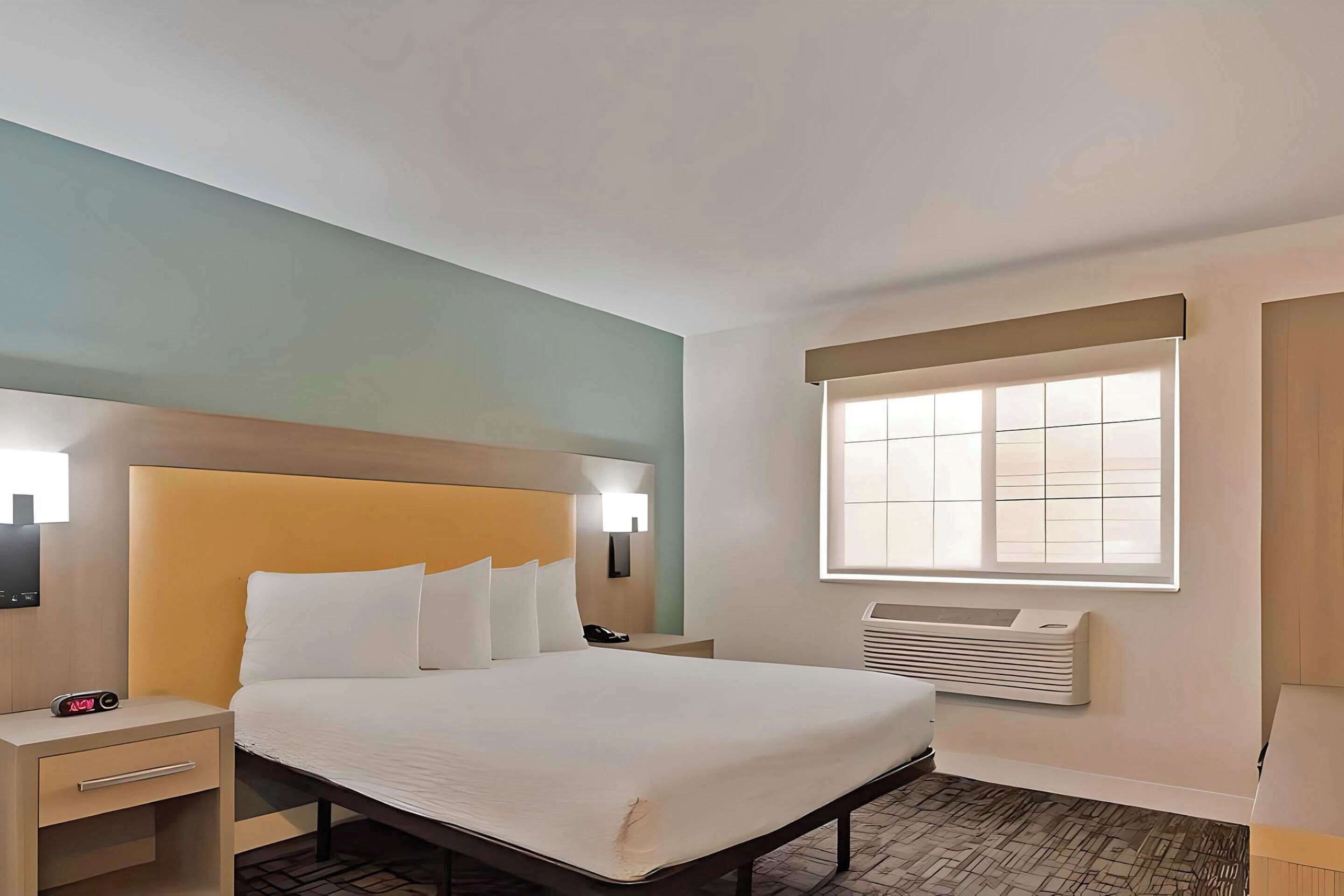 Une chambre d’hôtel comprenant un lit, des tables de chevet, une petite table, un radiateur fixé au mur et une fenêtre avec des stores.