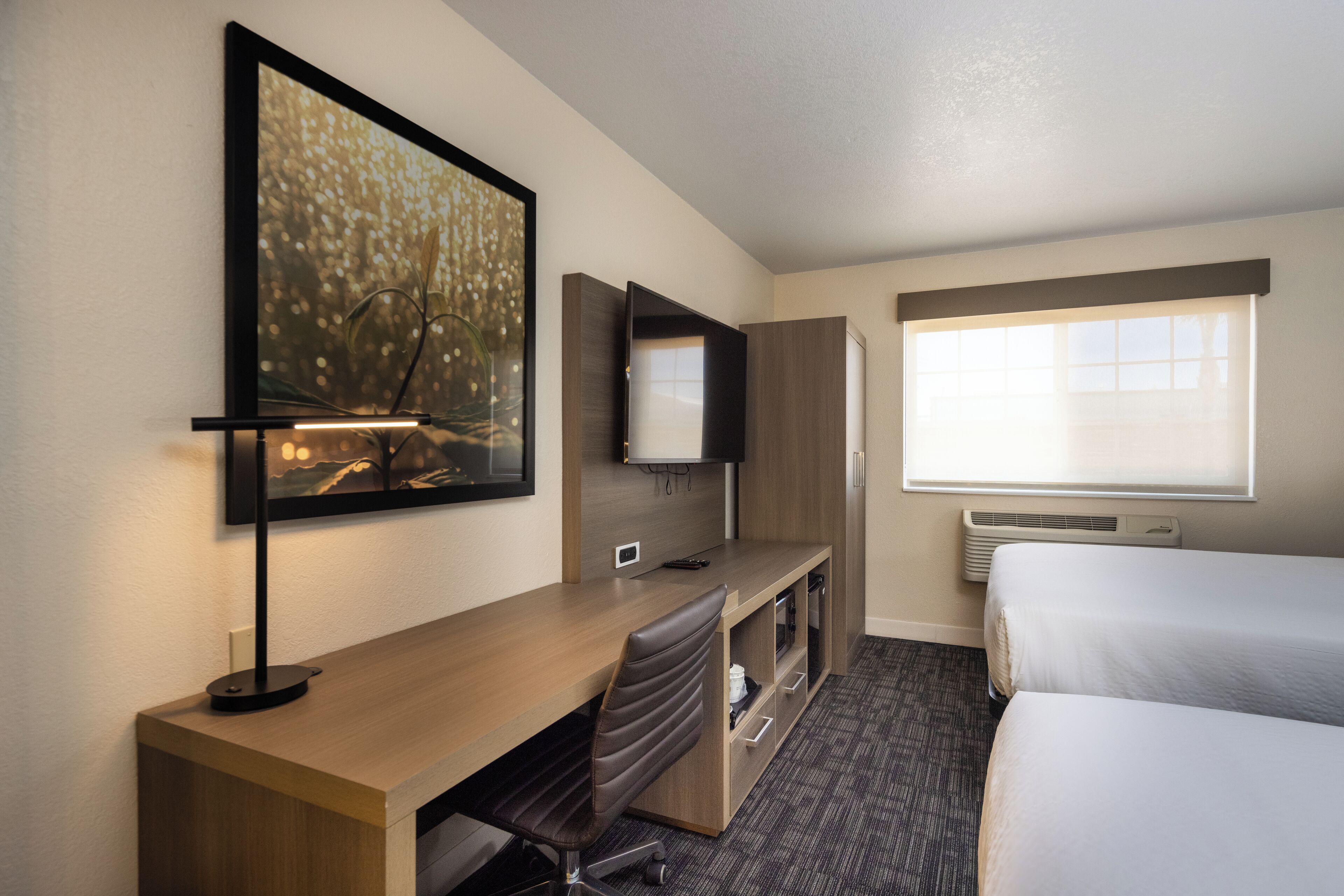 A hotel room with a desk, chair, bed, TV, and artwork on the wall.