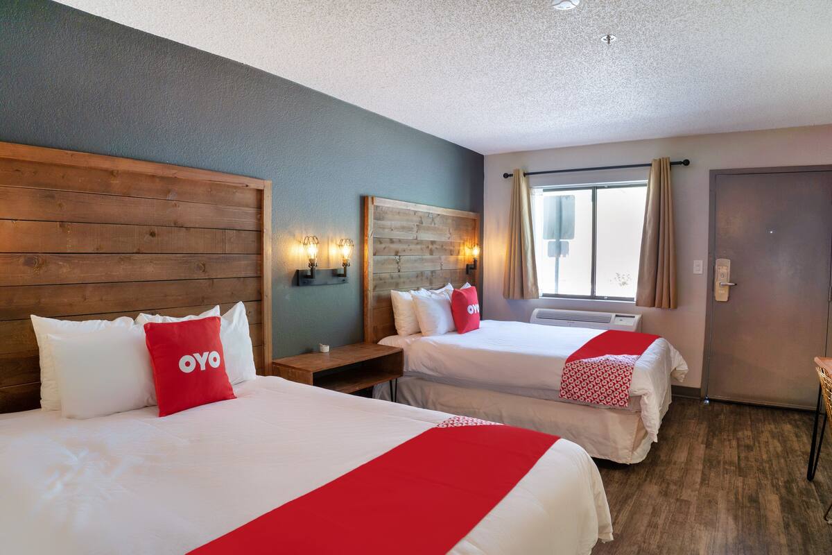 A hotel room with two beds, wooden headboard, bedside tables with lamps, a large window with curtains, and a door.