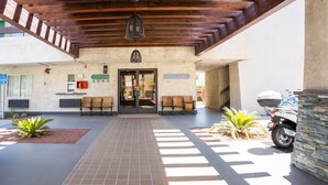 Un espace extérieur abrité, doté d’un sol carrelé, qui mène à l’entrée d’un bâtiment. Une moto est garée sur le côté, et un panneau indique la présence d’un club de plage.