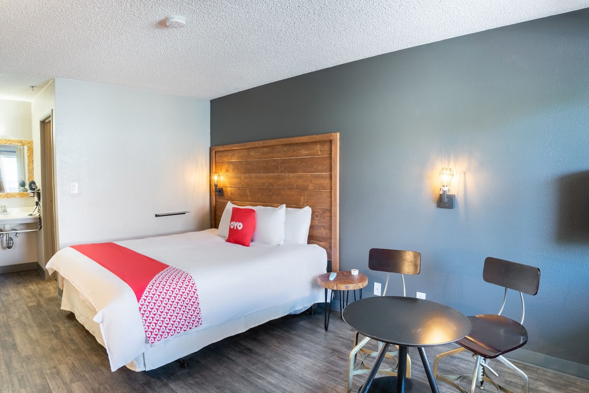 A hotel room with a large bed, a wooden headboard, a small round table with two chairs, and a wall-mounted lamp.