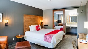 Une chambre d’hôtel avec un grand lit, une tête de lit en bois, une petite table ronde, un fauteuil en cuir, un banc en bois, un bureau avec une chaise et une salle de bain avec un miroir et un lavabo.