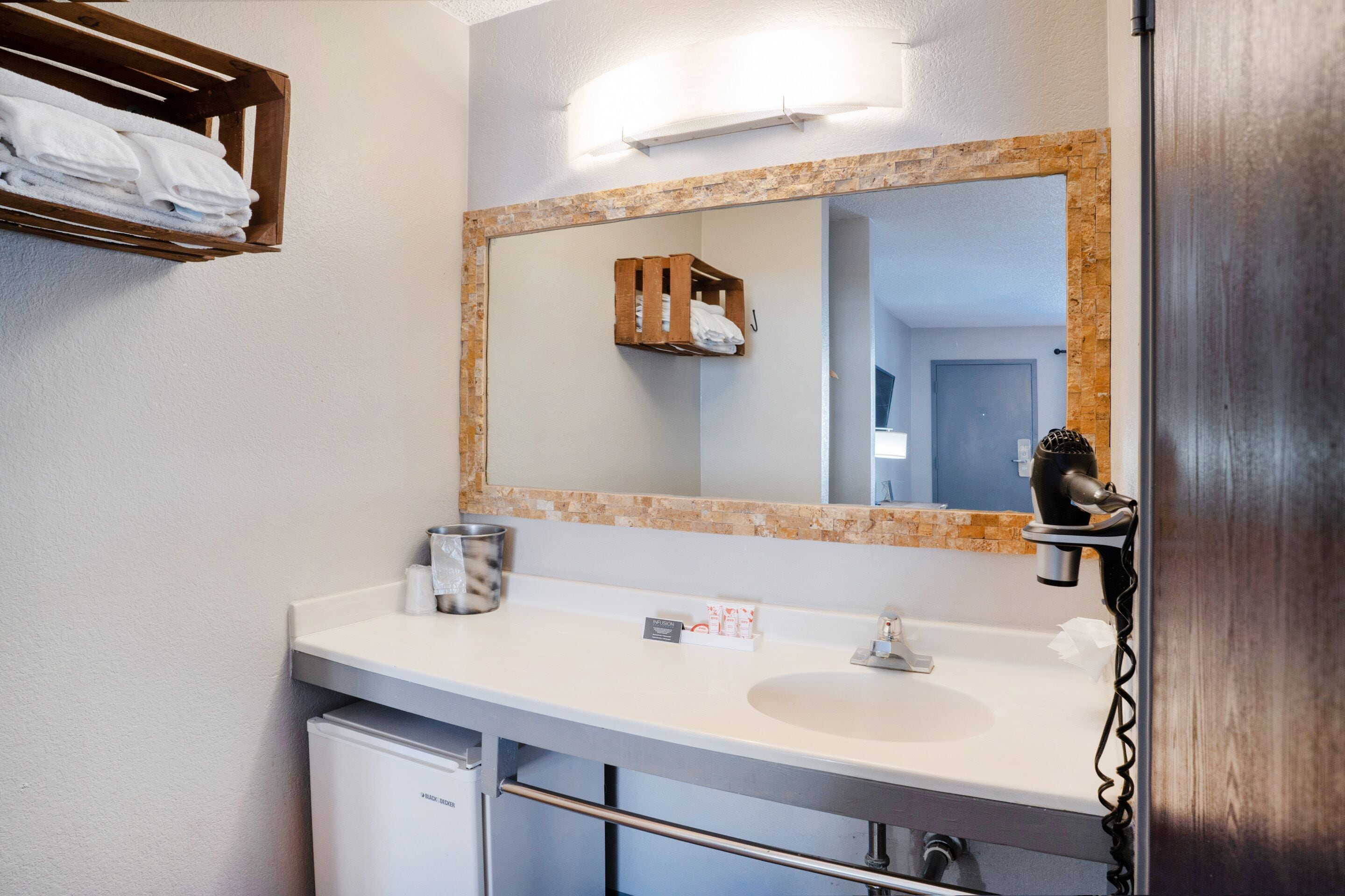 A bathroom with a white sink, a mirror with a wooden frame, a towel rack with folded towels, a hairdryer, and a small trash bin.
