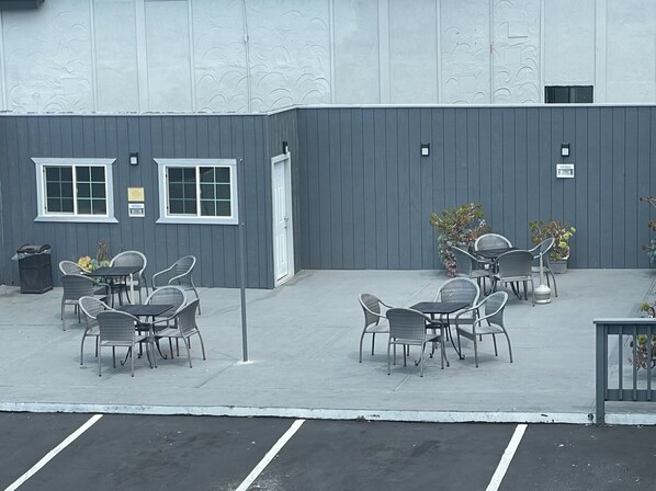 Un patio al aire libre con mesas y sillas, un edificio de fachada gris y un estacionamiento al frente.