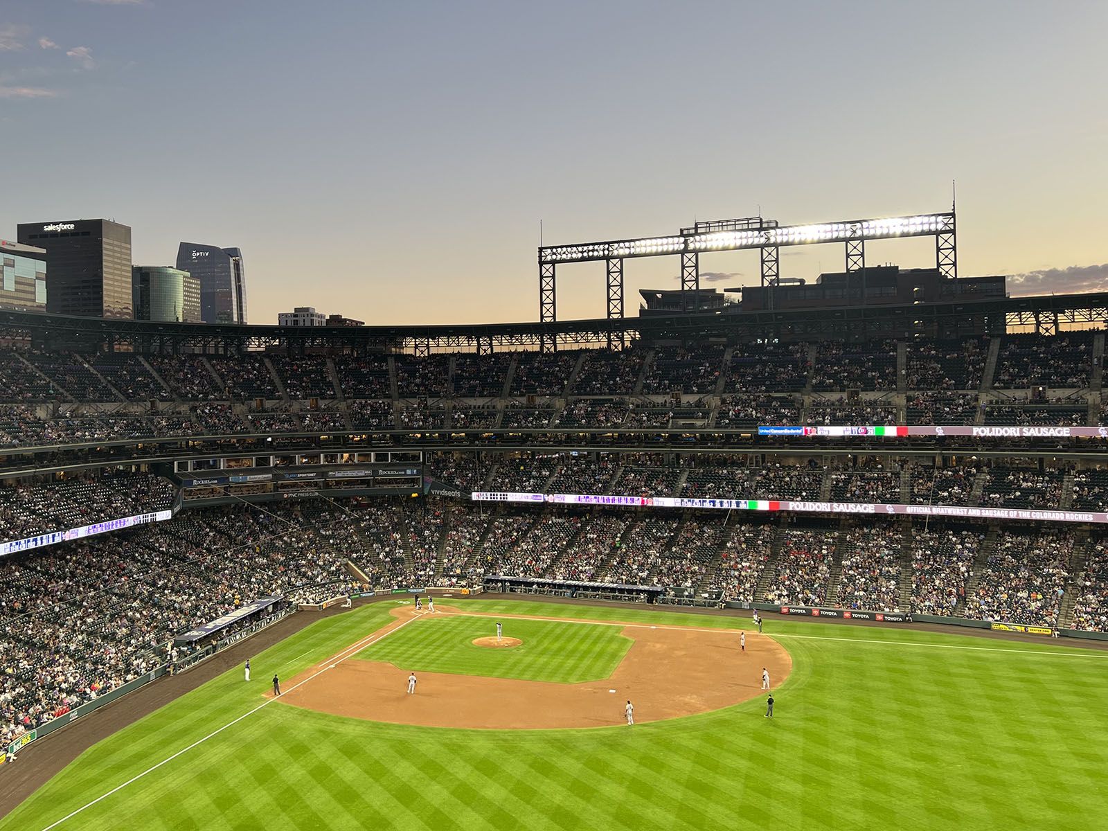 A baseball stadium with multiple tiers of seating, a large outfield, and a city skyline in the background.