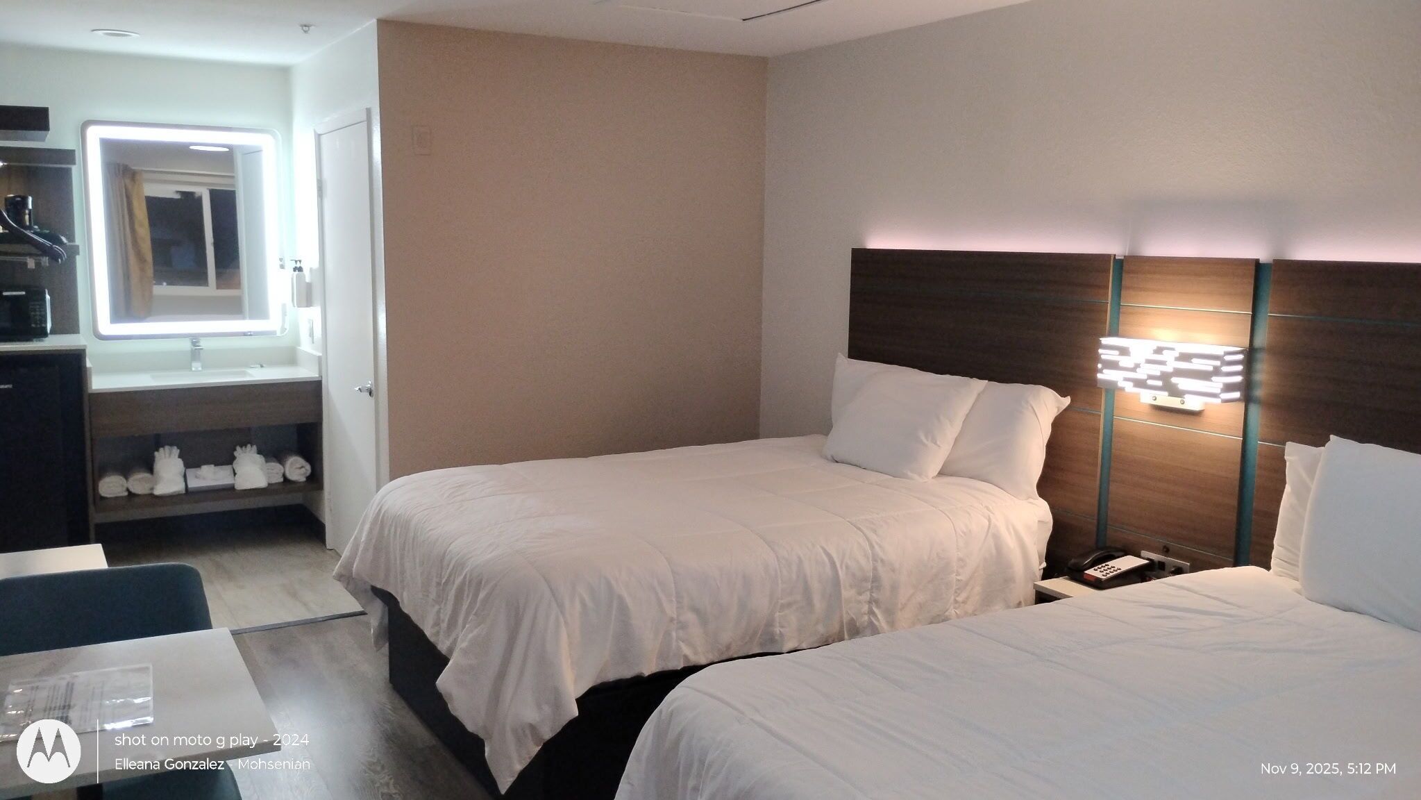A hotel room with two beds, a bedside table, a nightstand, a desk with a chair, a mirror, and a shelf with towels.
