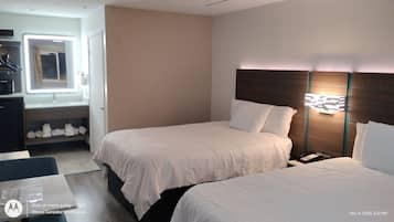 A hotel room with two beds, a bedside table, a nightstand, a desk with a chair, a mirror, and a shelf with towels.