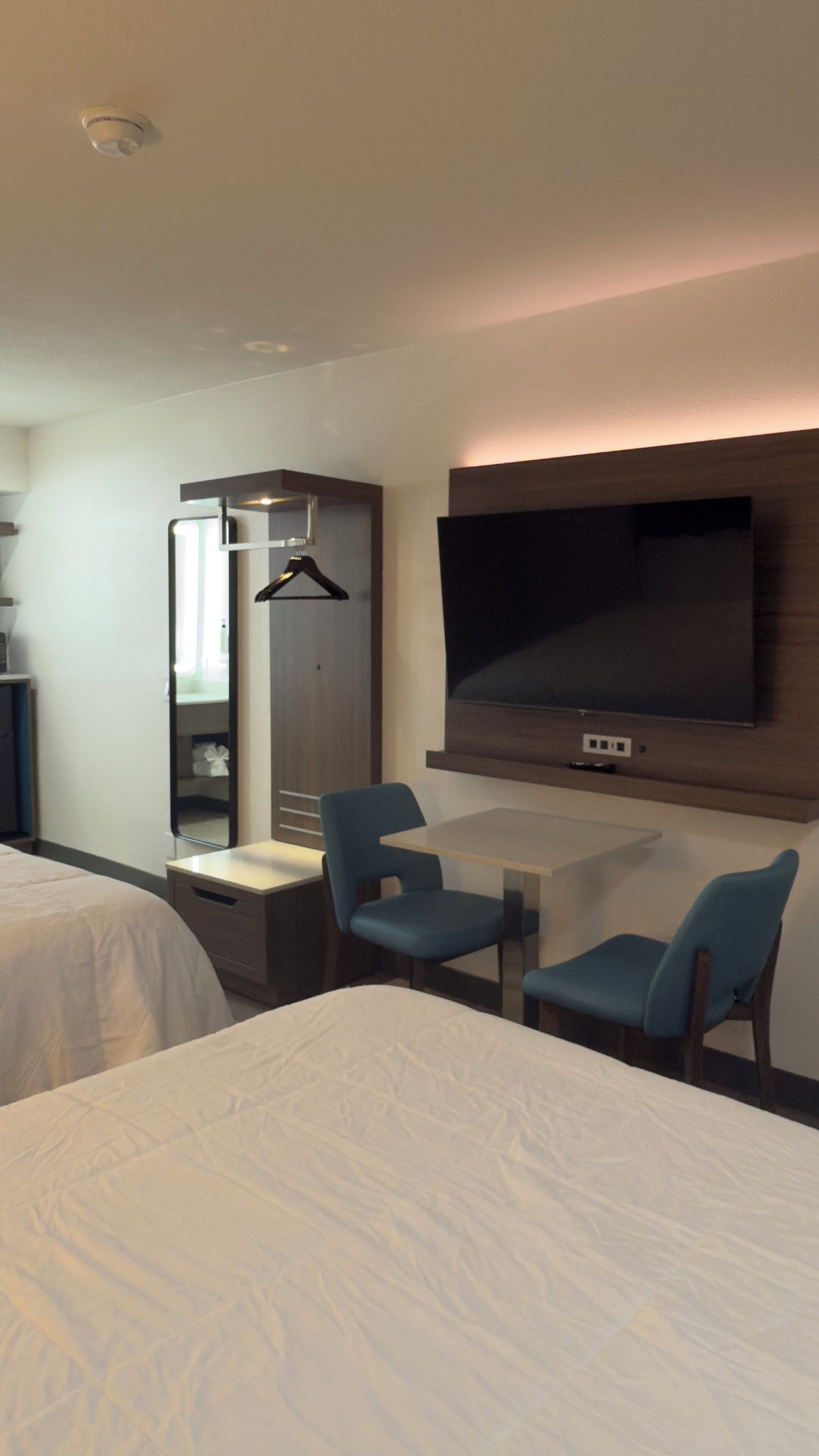 Habitación de hotel con cama, escritorio con dos sillas, televisor montado en la pared y armario.