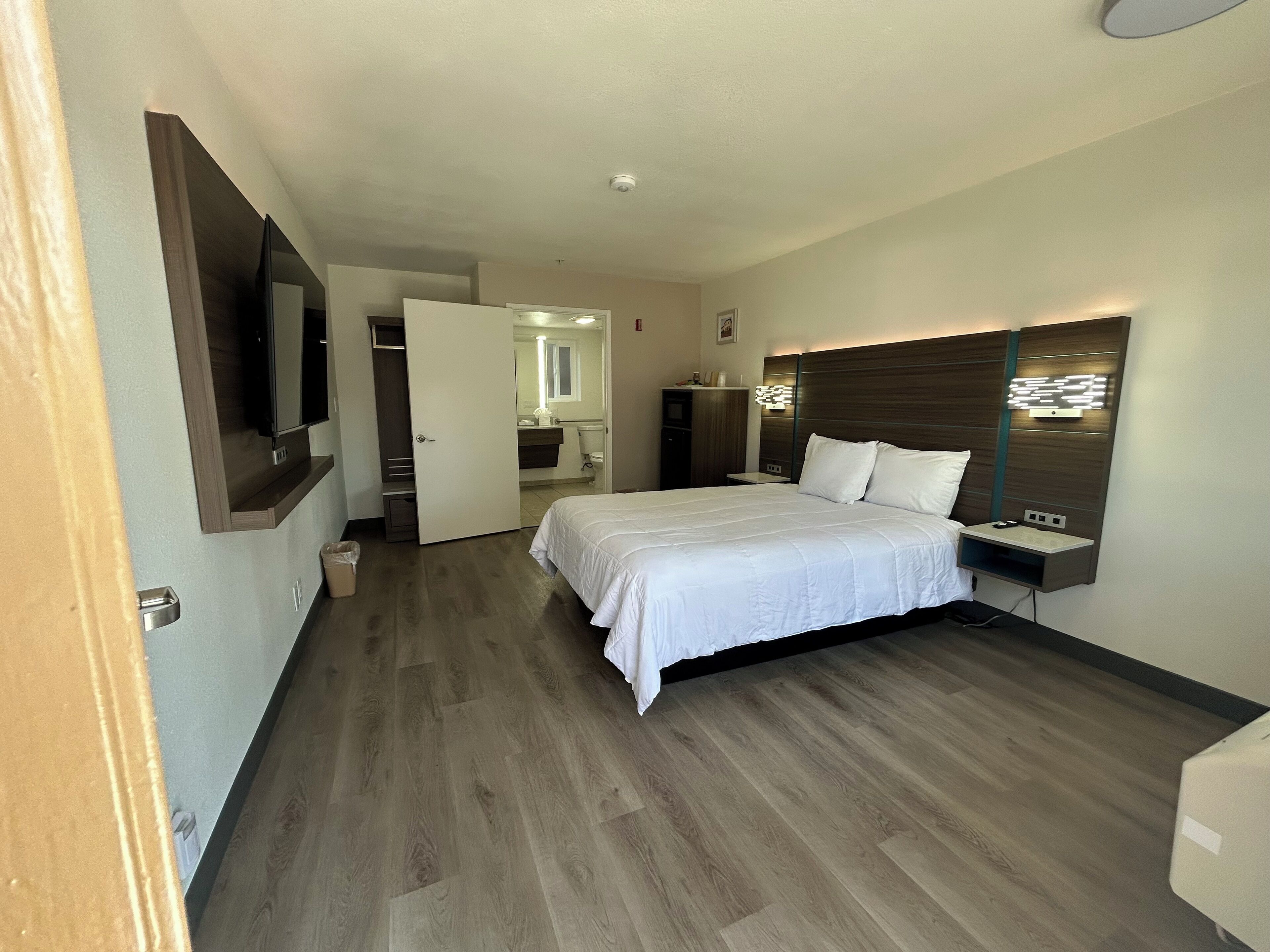A hotel room with a large bed, a bedside table, a TV mounted on the wall, and a bathroom visible through an open door.