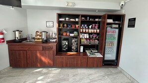 A compact kitchen with a coffee machine, a trash bin, a refrigerator stocked with beverages, and shelves with snacks and condiments. - Mission Inn San Luis Obispo (San Luis Obispo)