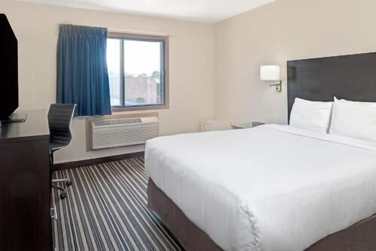 Una habitación de hotel con cama, escritorio, silla, ventana con cortinas, lámpara de pared y radiador.