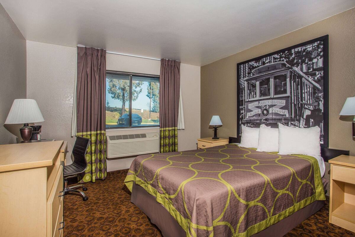 A hotel room with a bed, a desk, a chair, two lamps, a window with curtains, and a wall with a black and white photo.