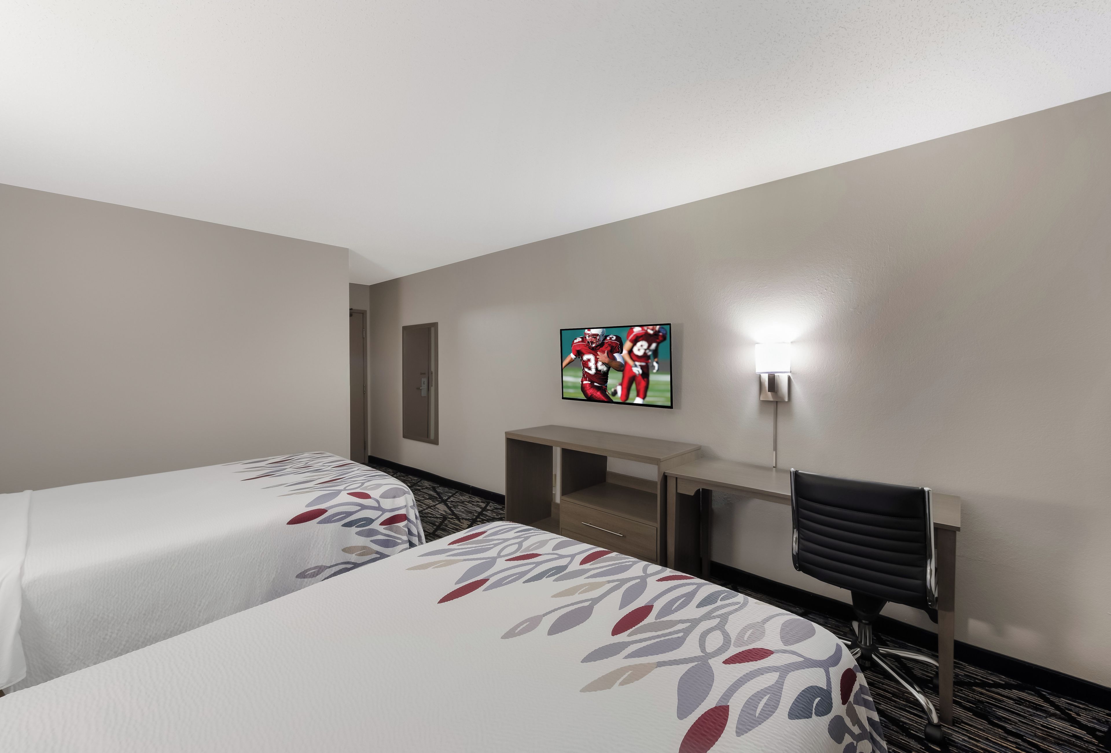 A hotel room with two beds, a desk, a chair, a TV, and a wall-mounted light.