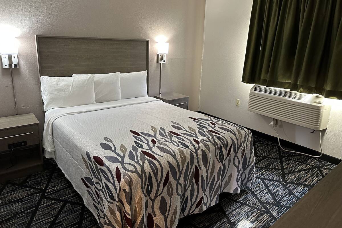 A hotel room with a bed, bedside tables, a wall-mounted air conditioner, and a window with curtains.