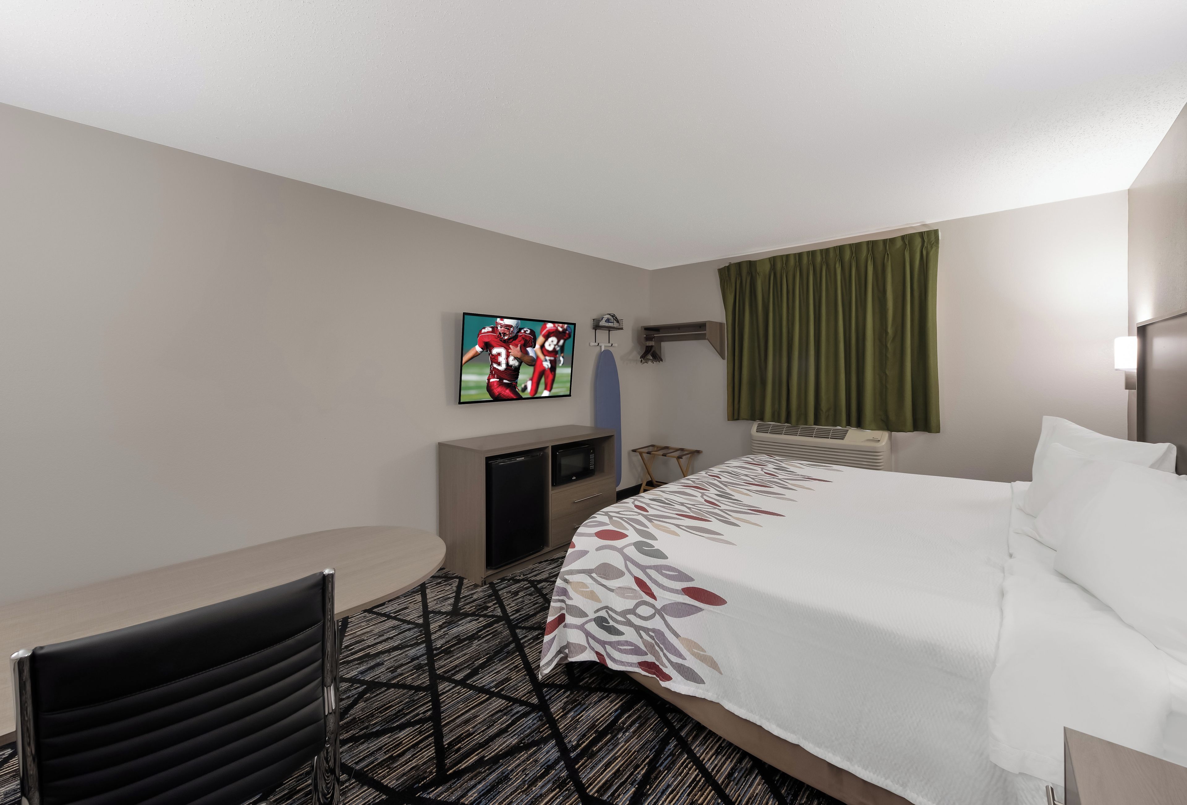 Una camera d'albergo con un letto, una scrivania, un televisore, un piccolo frigorifero e una finestra con tende.
