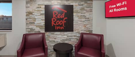 Red Roof Inn -hotellin huone, jossa on kiviseinä, kaksi nahkaista nojatuolia, pieni pöytä ja kyltti, joka ilmoittaa, että kaikissa huoneissa on käytettävissä ilmainen Wi-Fi-yhteys.