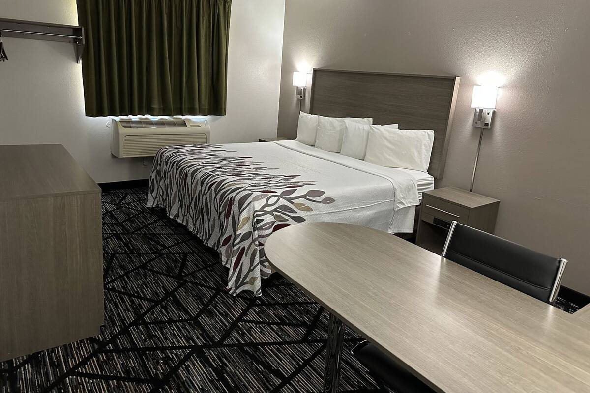 A hotel room with a bed, a desk, a chair, a lamp, and a window with curtains.
