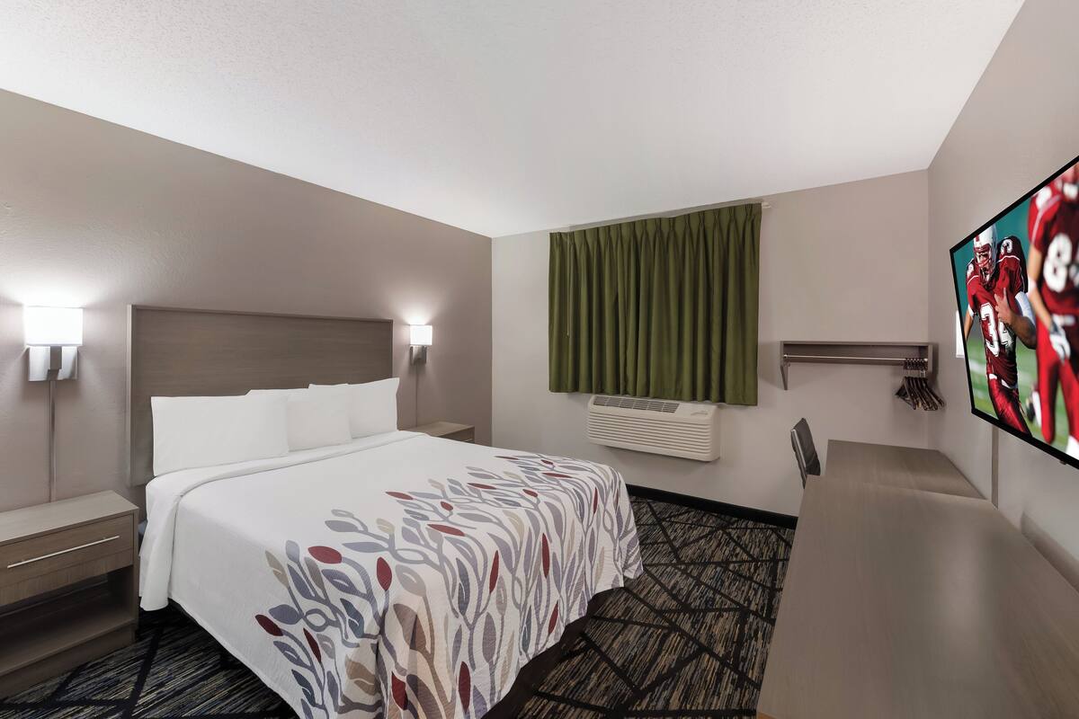 A hotel room with a bed, bedside lamps, a flat-screen TV, an air conditioning unit, and a green curtain.