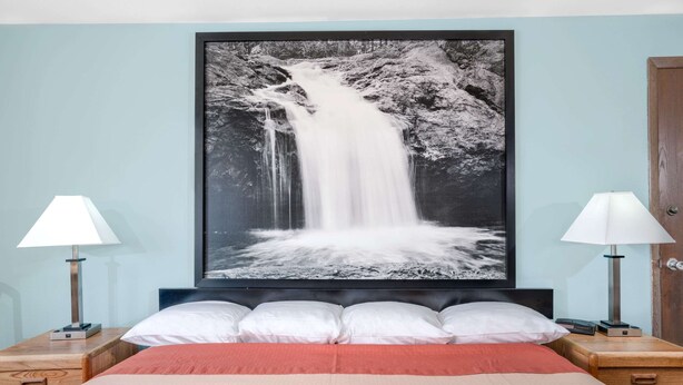 A bed with a red blanket, white pillows, and a black headboard. There is a framed black and white waterfall painting above the bed. Two bedside tables with lamps are on either side of the bed.