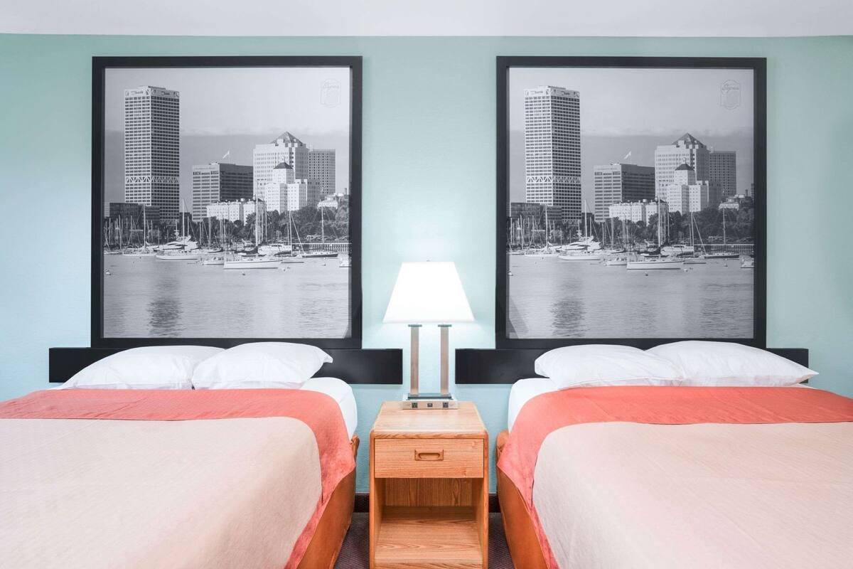 A hotel room with two beds, a nightstand with a lamp, and two framed cityscape photographs on the wall.