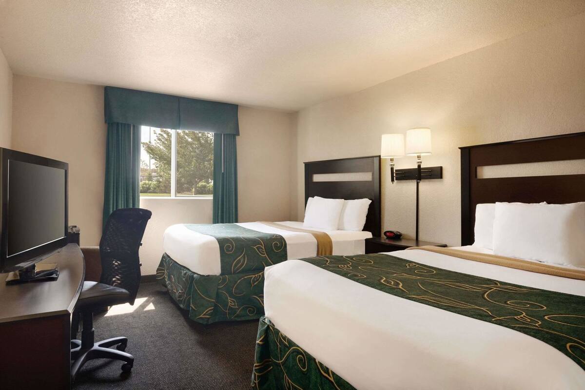Habitación de hotel con dos camas, un escritorio con televisor, una silla, una ventana con cortinas y una mesita de noche con lámparas.