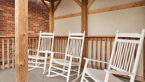 Three white rocking chairs in a covered outdoor area with exposed wooden beams and a brick wall.