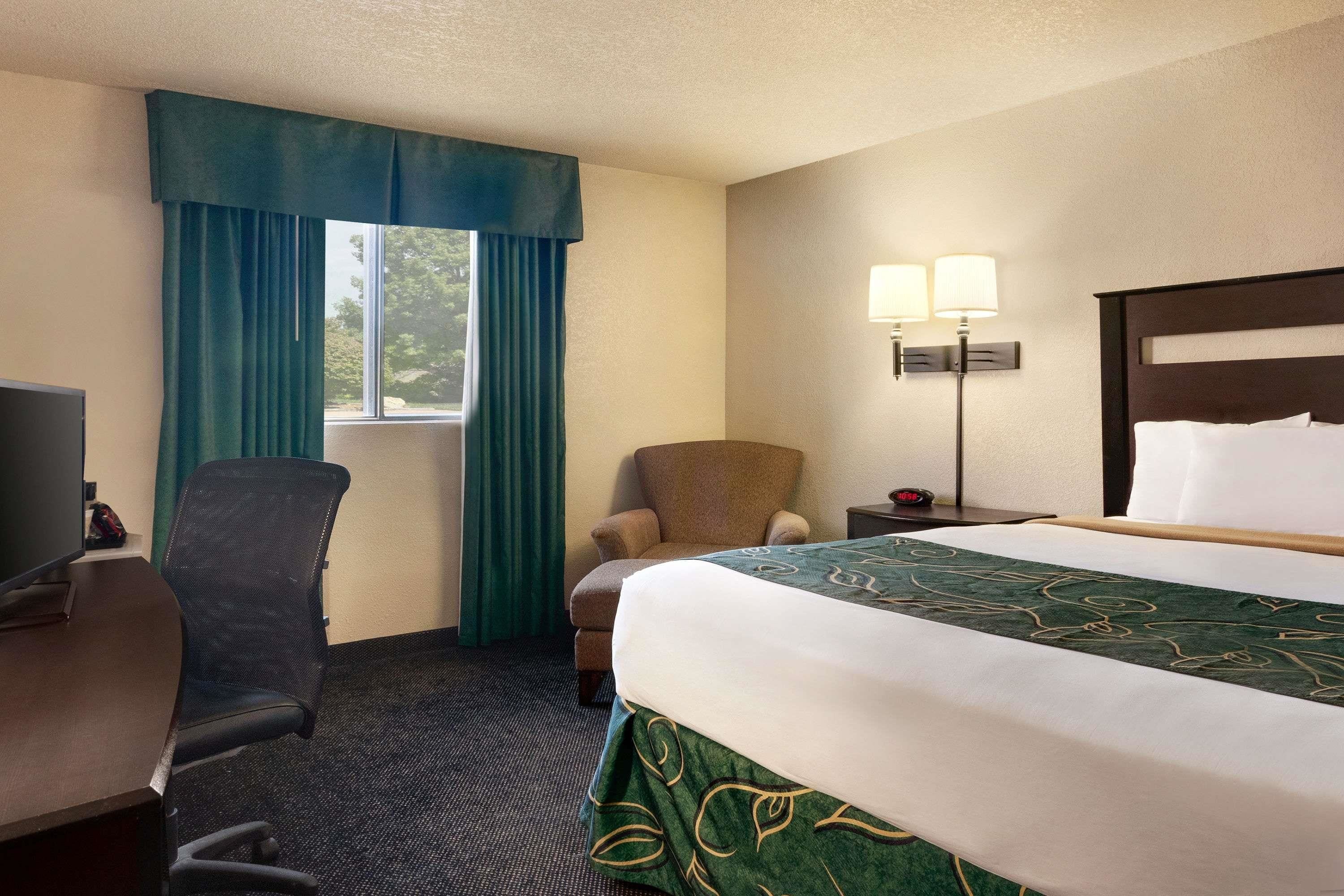 Habitación de hotel con cama, escritorio, silla, lámpara y ventana con cortinas.