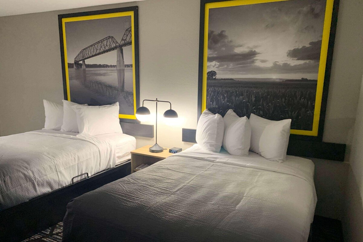 A hotel room with two beds, a nightstand, a lamp, and two framed pictures on the wall.