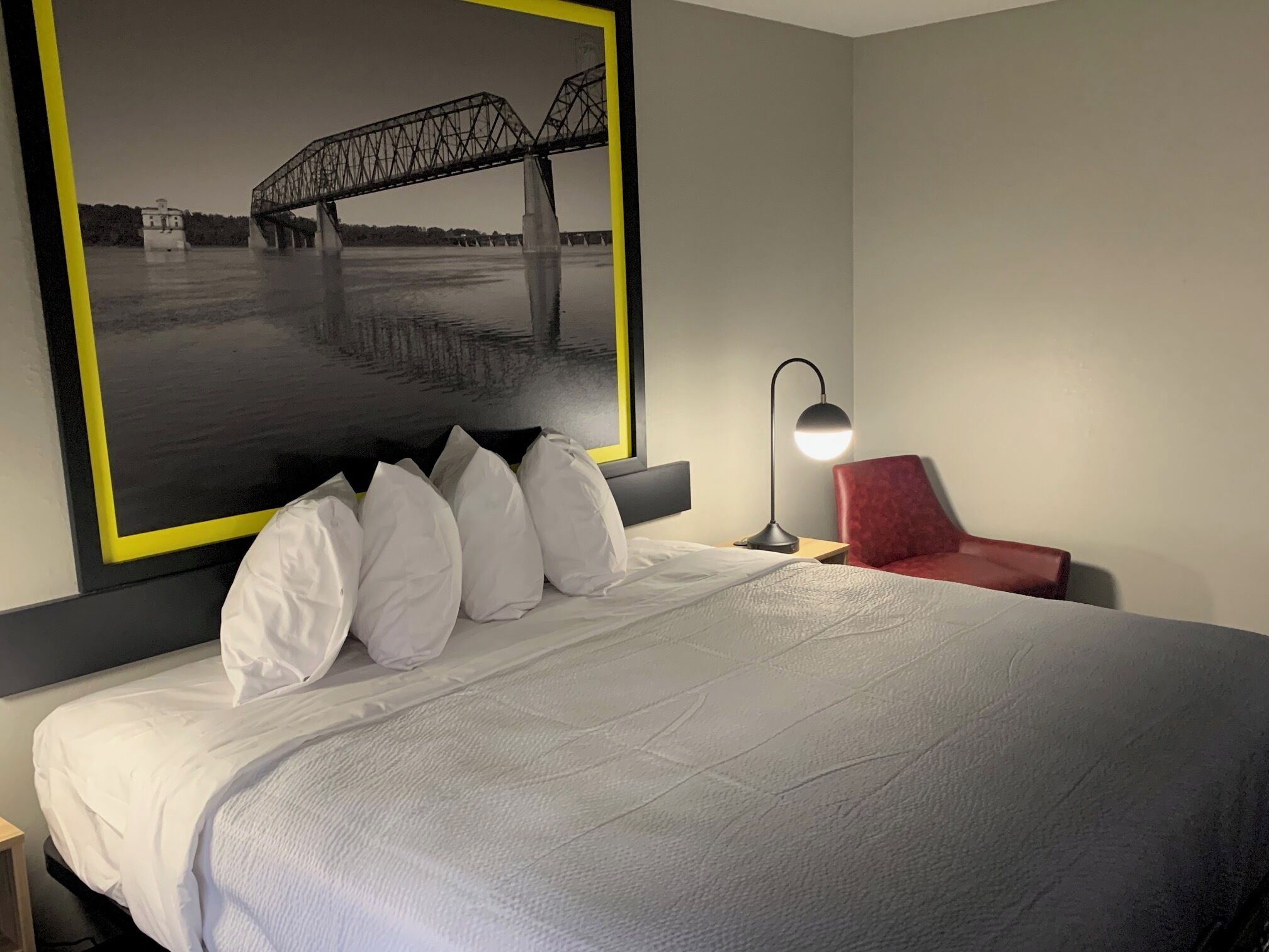 Ett hotellrum med en stor säng, en röd stol, ett skrivbord med en lampa och en inramad bild av en bro.