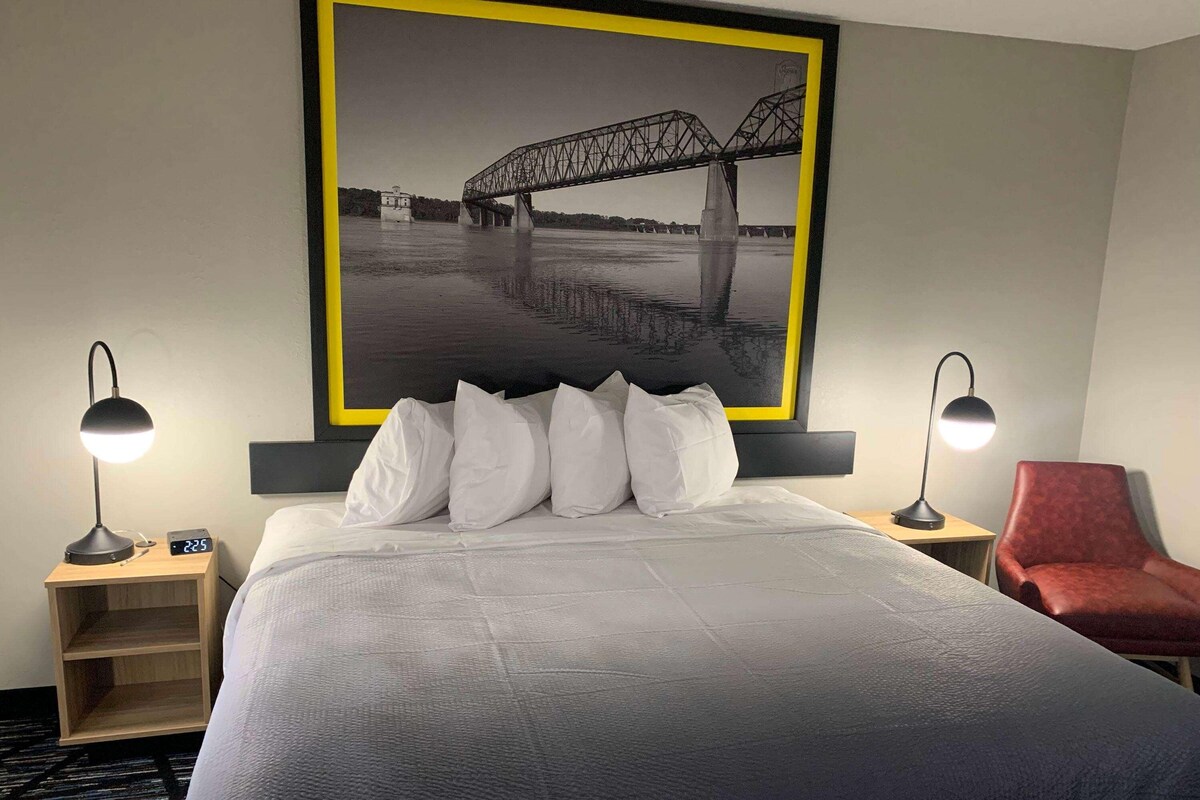 A hotel room with a large bed, two bedside lamps, a red chair, and a framed black and white photo of a bridge.