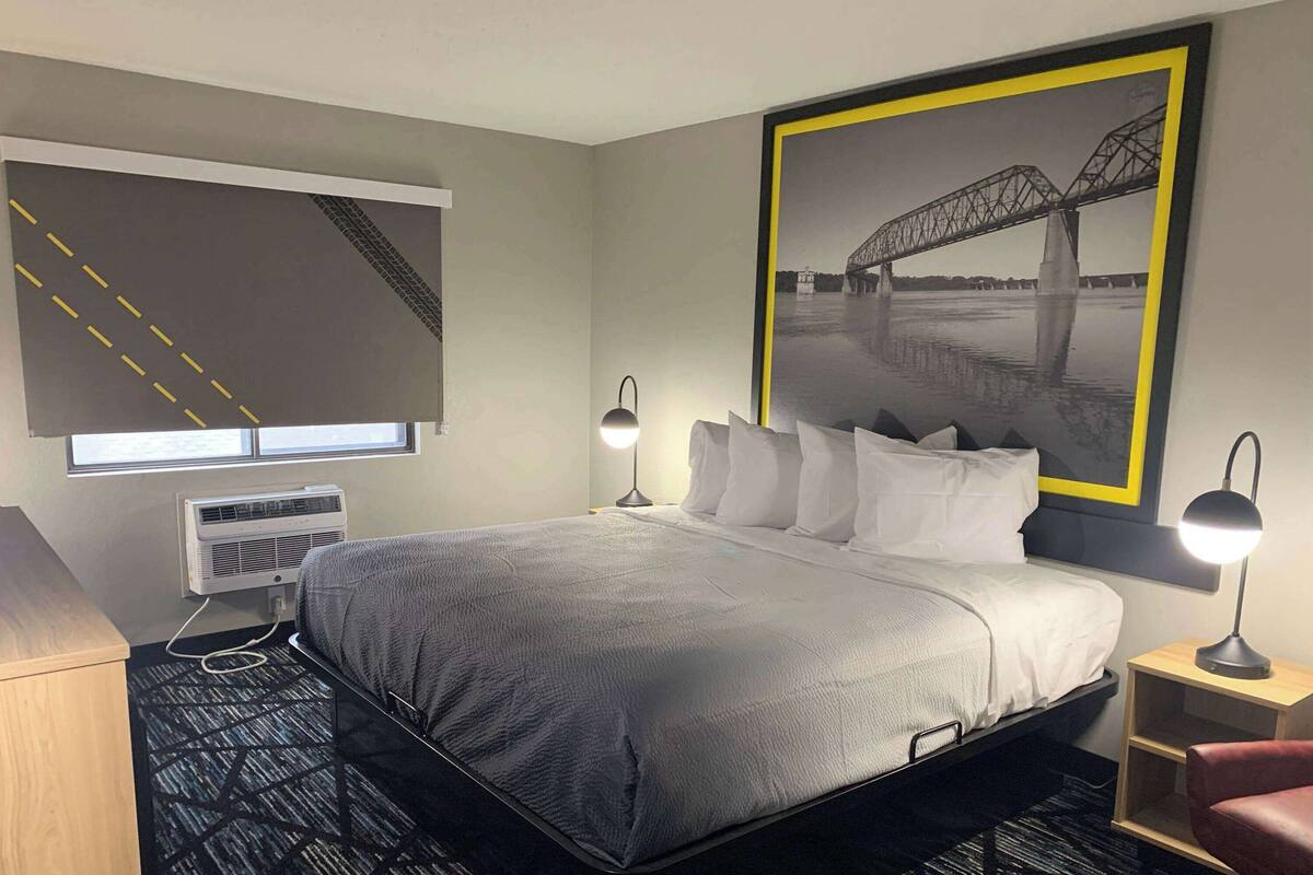 A hotel room with a bed, bedside lamps, a window with blinds, an air conditioner, a desk, and a framed picture on the wall.
