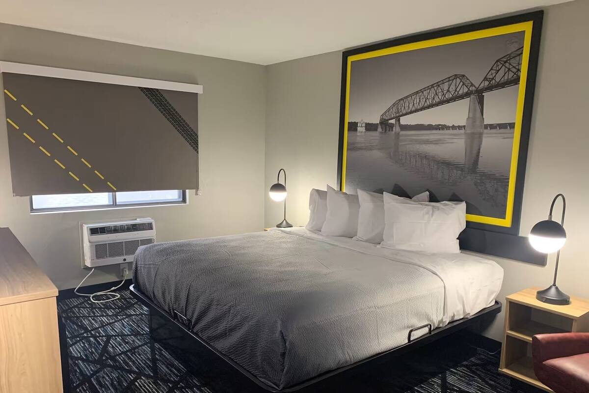 A hotel room with a bed, bedside lamps, a window with blinds, an air conditioning unit, a desk, and a chair.
