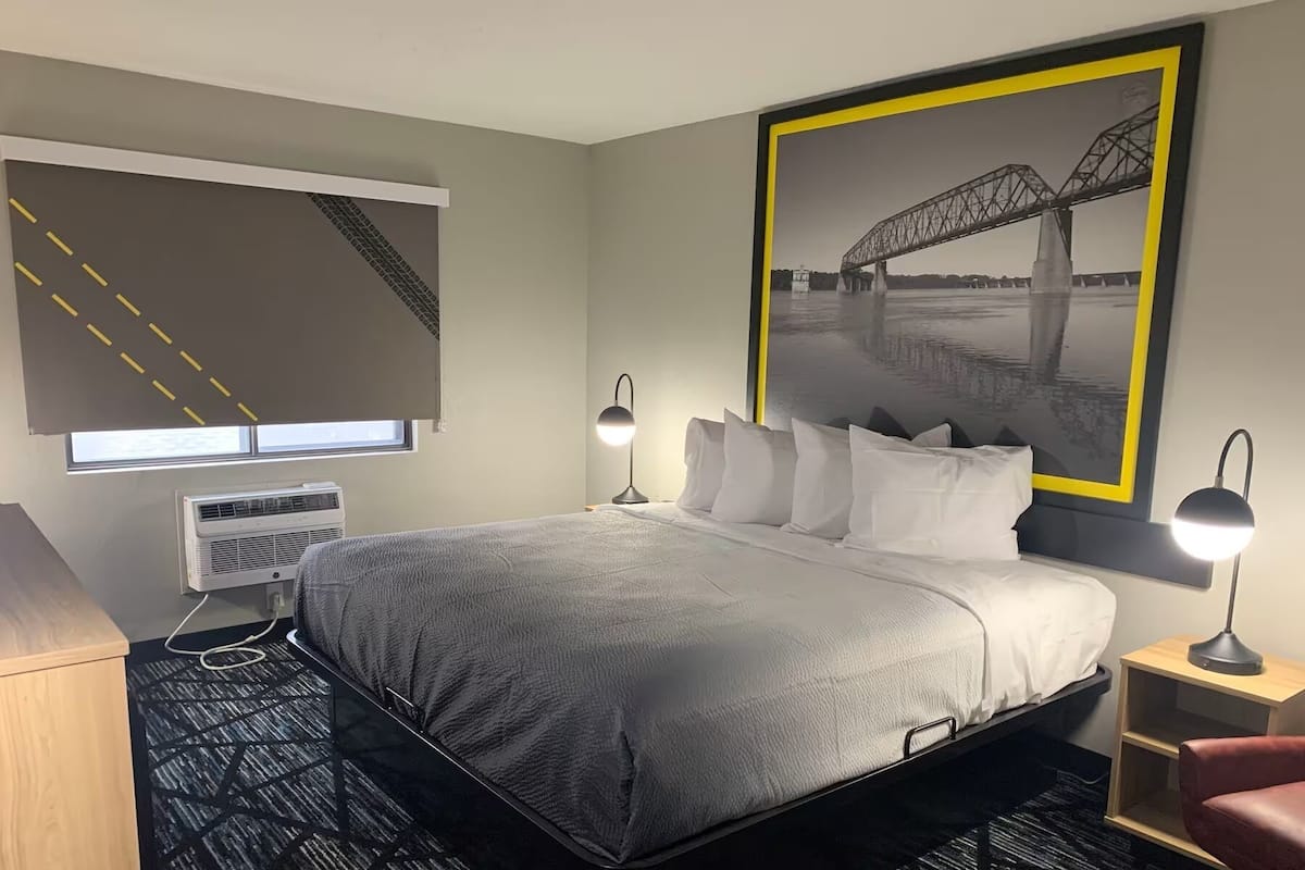 Une chambre d’hôtel comprenant un lit, des lampes de chevet, une fenêtre avec des stores, un climatiseur, un bureau et une chaise.