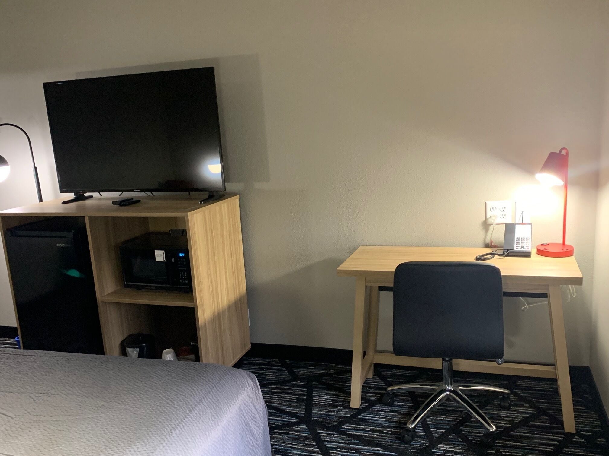A hotel room with a bed, a desk with a chair, a TV, and a lamp.